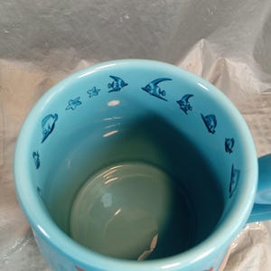 May include: A light blue ceramic mug featuring a cartoon mermaid design. The mug's interior is a lighter shade of blue, with small fish and star illustrations around the rim. The handle is also light blue.
