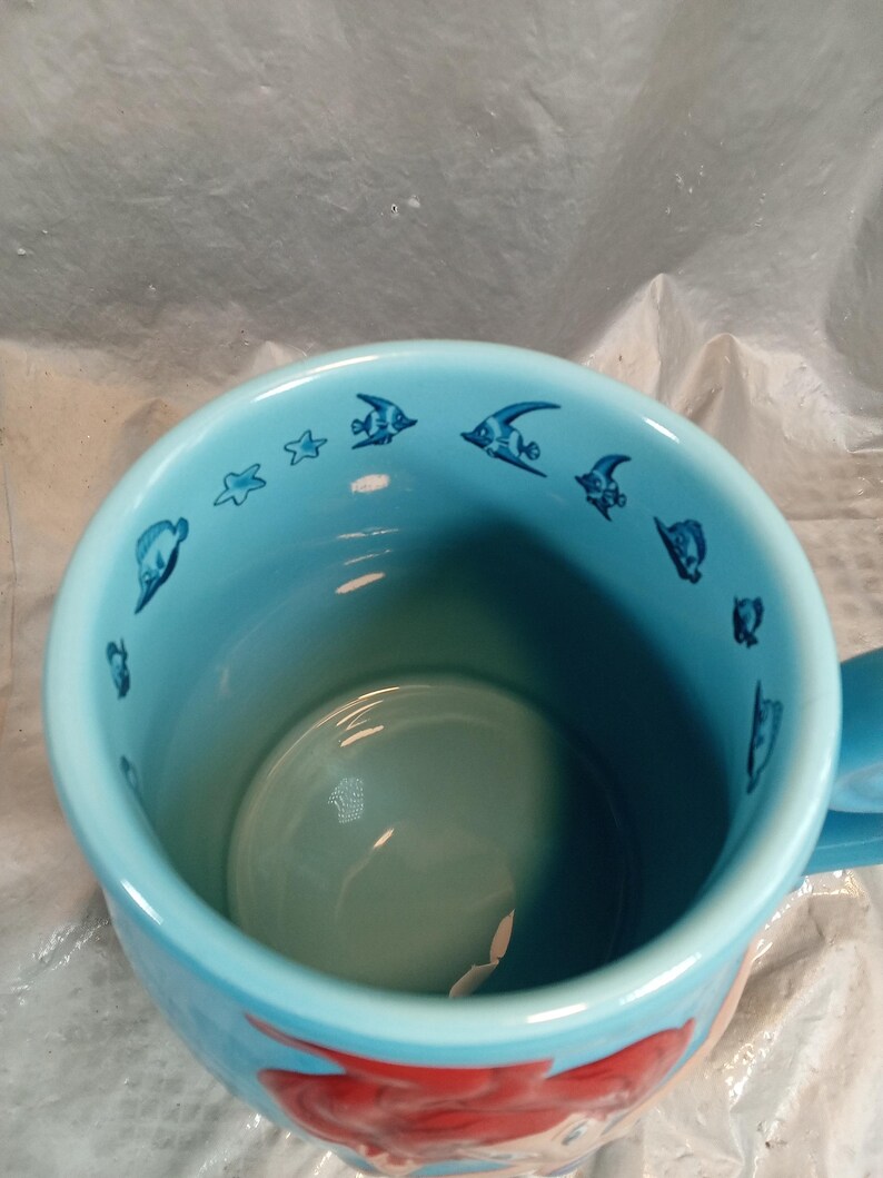 May include: A light blue ceramic mug featuring a cartoon mermaid design. The mug's interior is a lighter shade of blue, with small fish and star illustrations around the rim. The handle is also light blue.