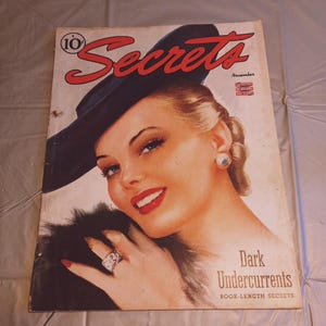 Puede incluir: Una portada de revista vintage con una mujer que lleva un sombrero negro y un cuello de piel. La revista se titula "Secrets" y la historia de portada es "Dark Undercurrents, Book-Length Secrets".