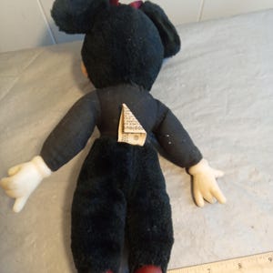 May include: A vintage Minnie Mouse plush doll, viewed from the back. The doll has a black body, white gloves, and red shoes. A red bow adorns the head. A tag is attached to the back. The doll is placed on a surface with a ruler.