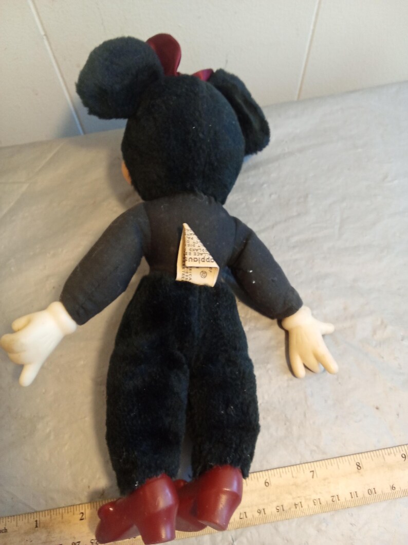 May include: A vintage Minnie Mouse plush doll, viewed from the back. The doll has a black body, white gloves, and red shoes. A red bow adorns the head. A tag is attached to the back. The doll is placed on a surface with a ruler.