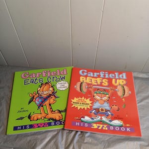 May include: Two Garfield comic books, one green with the title "Garfield Eats Crow" and the other red with the title "Garfield Reefs Up". Both books feature Garfield the cat in a cartoon illustration.