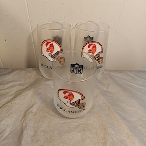 Könnte beinhalten: Drei transparente Trinkgläser mit dem Logo der Tampa Bay Buccaneers. Das Logo zeigt einen rot-orangen Helm mit dem Teamnamen "Buccaneers" darunter. Das NFL-Logo ist ebenfalls sichtbar.