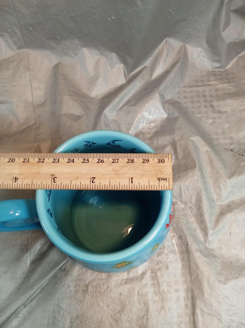 May include: A light blue ceramic mug with a handle, featuring a sea life design. The mug has a diameter of approximately 4 inches. The interior is green, and the rim is decorated with fish illustrations.