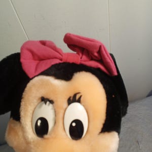 May include: A plush toy featuring a cartoon character with a black head, large white eyes, and a pink bow. The character has a tan face and a black nose. The toy is wearing a green shirt.