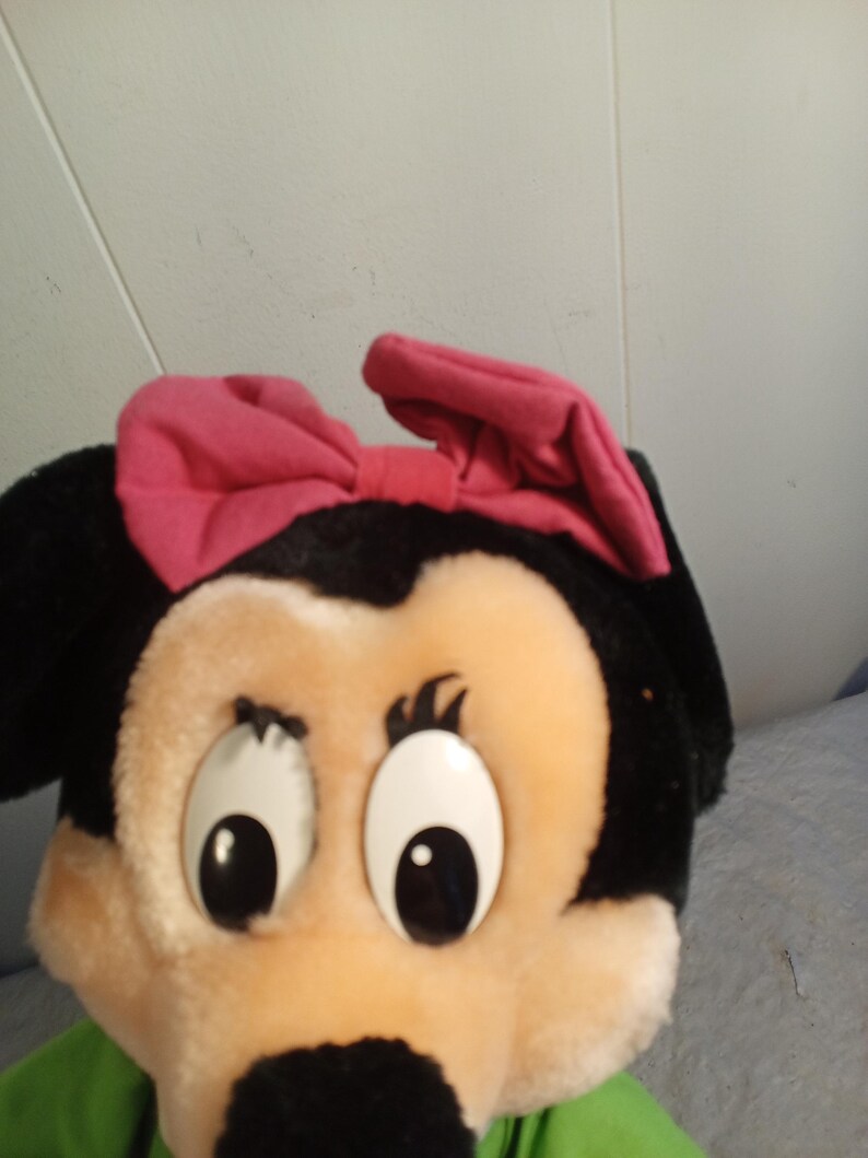 May include: A plush toy featuring a cartoon character with a black head, large white eyes, and a pink bow. The character has a tan face and a black nose. The toy is wearing a green shirt.