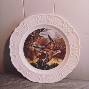 May include: White decorative plate with a scalloped edge and embossed floral design. The center features a detailed painting of two birds in flight over a landscape with brown and green tones. The plate is likely ceramic.