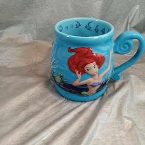 May include: A light blue ceramic mug featuring Ariel from The Little Mermaid. The mug has a curved handle and is decorated with images of fish, sea turtles, and Ariel. The mug is a collectible item.