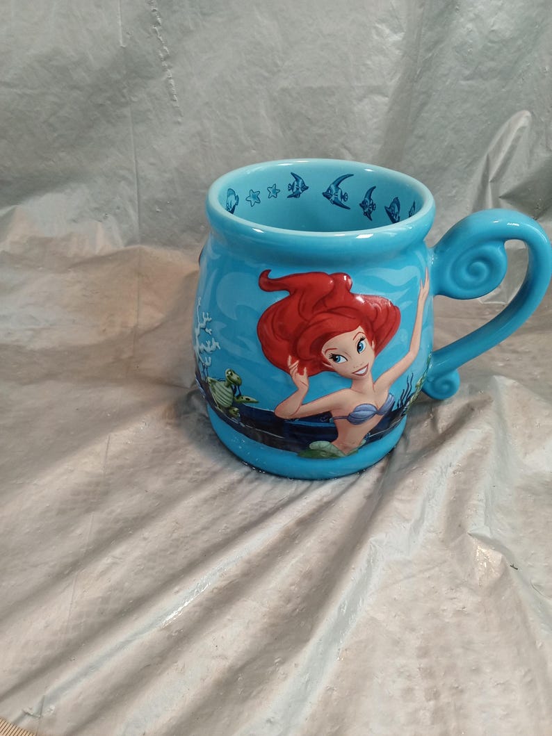 May include: A light blue ceramic mug featuring Ariel from The Little Mermaid. The mug has a curved handle and is decorated with images of fish, sea turtles, and Ariel. The mug is a collectible item.