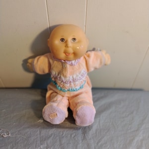 Può includere: Una bambola Cabbage Patch Kid calva che indossa un pigiama rosa e bianco con un bordo blu. La bambola ha gli occhi marroni e un grande sorriso.