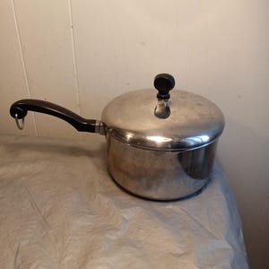 May include: A stainless steel saucepan with a lid and a black handle. The pot has a black knob on the lid and a metal loop at the end of the handle. The pot is sitting on a wrinkled, light-colored surface.