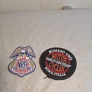 Op de afbeelding: Twee geborduurde patches. Eén is een schildvorm met een adelaar, de tekst "LAW NRA ENFORCEMENT". De andere is een zwarte cirkel met "NUMERO UNO MOTOR HARLEY-DAVIDSON CYCLES ROMA ITALIA" in oranje en wit.