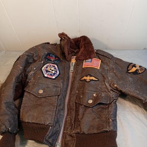 May include: Brown bomber jacket with patches, a fur collar, and a zipper closure. The jacket features an American flag patch and other military-themed patches. The jacket has two front pockets and ribbed cuffs and hem.