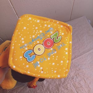 Puede incluir: Un juguete de peluche amarillo con una etiqueta cuadrada con el texto "2003 The One To Be" y "Walt Disney World" en azul, rojo y verde. La etiqueta tiene un patr&oacute;n de estrellas blancas.
