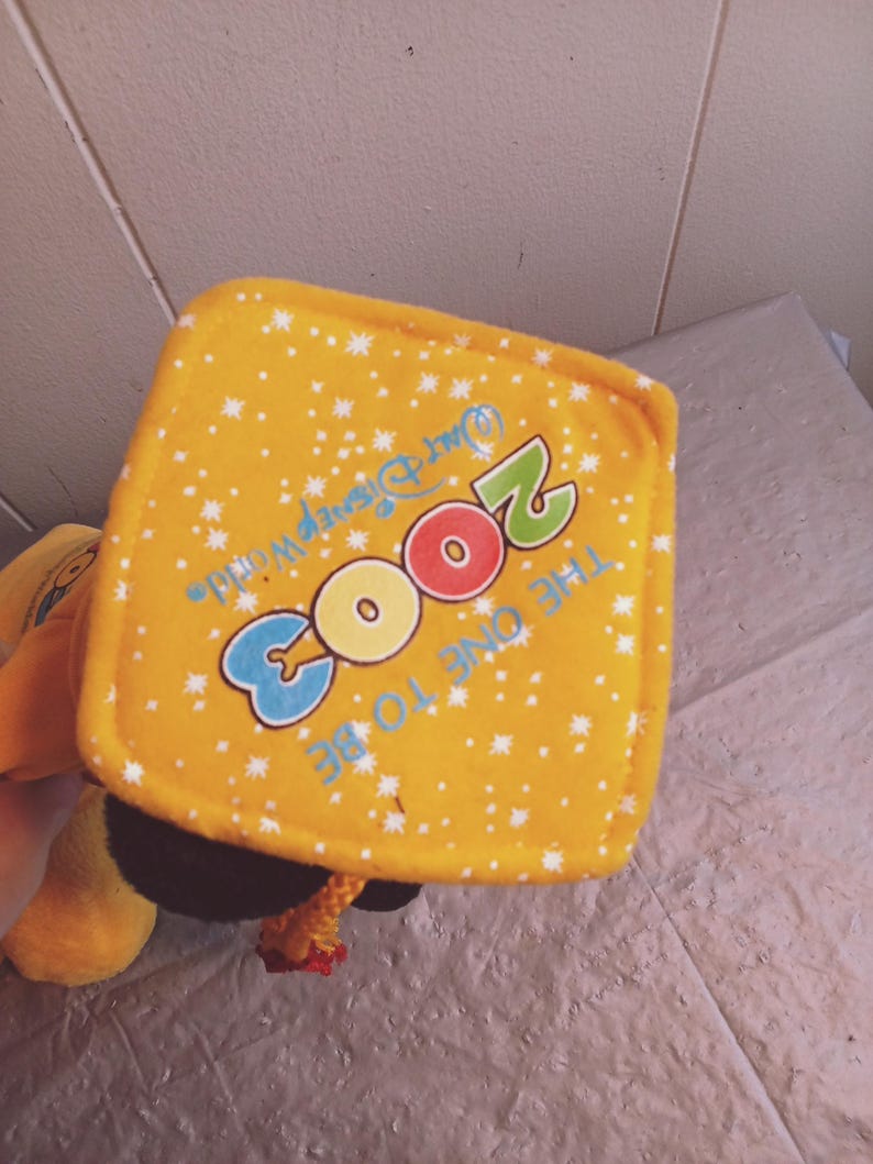 Puede incluir: Un juguete de peluche amarillo con una etiqueta cuadrada con el texto "2003 The One To Be" y "Walt Disney World" en azul, rojo y verde. La etiqueta tiene un patr&oacute;n de estrellas blancas.