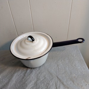 May include: White enamel saucepan with a lid and a dark blue handle. The rim and handle are outlined in black. The lid has a black handle. The saucepan is sitting on a gray surface.