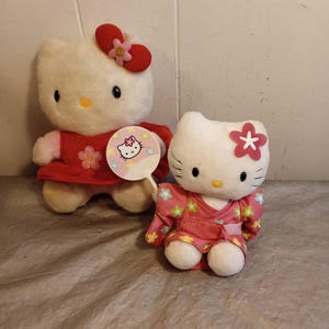 Puede incluir: Dos muñecos de peluche de Hello Kitty. Uno lleva un vestido rojo y un lazo, el otro un kimono rosa. Un abanico con un gráfico de Hello Kitty lo sostiene la primera muñeca. Ambas muñecas tienen ojos negros, nariz amarilla y pelaje blanco.