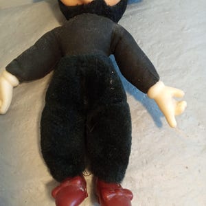 May include: A vintage Mickey Mouse doll. The doll has a black body, black ears, and a tan face. It is wearing a black shirt and pants, with red shoes. The doll's hands are white.