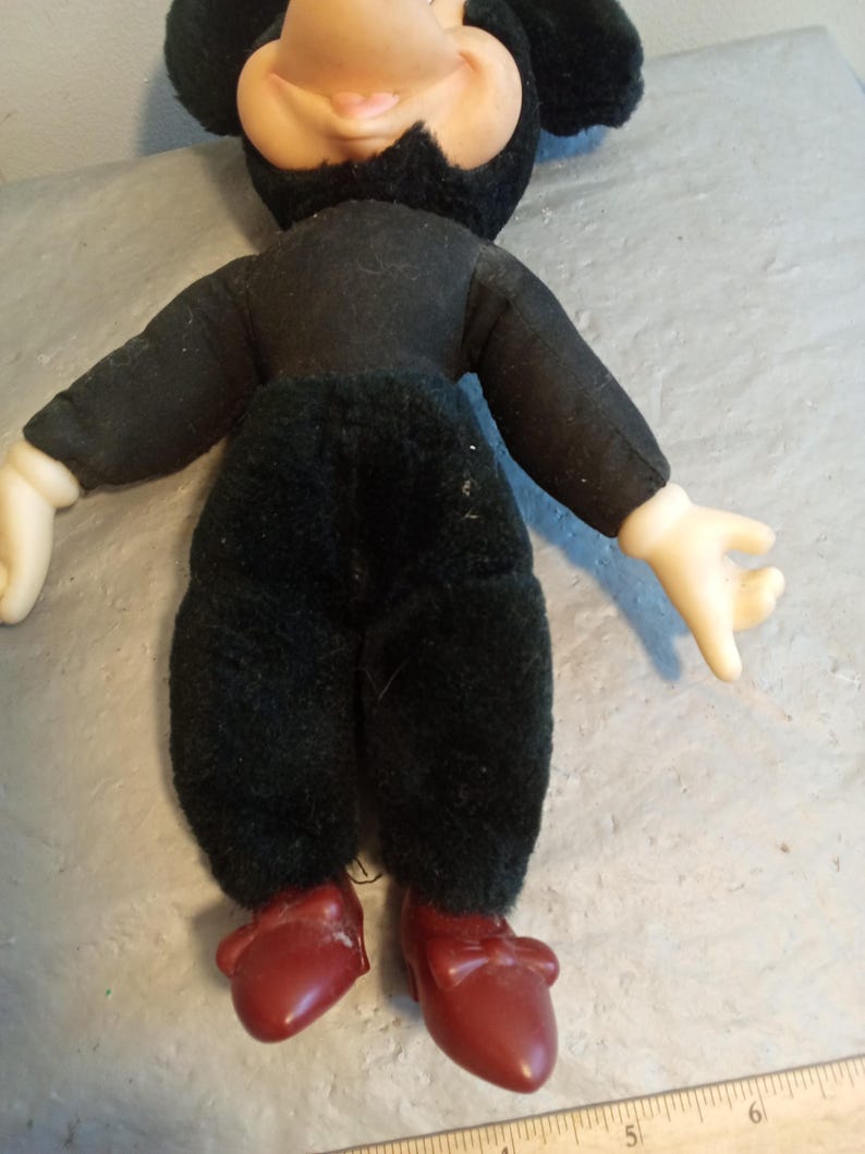 May include: A vintage Mickey Mouse doll. The doll has a black body, black ears, and a tan face. It is wearing a black shirt and pants, with red shoes. The doll's hands are white.