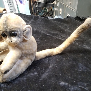May include: A vintage stuffed monkey with a light beige body, large black eyes, and a long, curved tail. The monkey is sitting on a dark surface, showcasing its detailed features and aged appearance. The toy's ears are large and rounded.