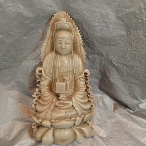 May include: A light beige stone sculpture of a seated figure with closed eyes, wearing flowing robes and holding a small rectangular object. The figure is positioned on a lotus-shaped base with an ornate, pointed, leaf-like halo behind the head.
