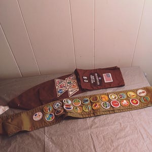 May include: A brown Boy Scouts sash with various merit badges and a brown neckerchief slide with merit badges. A brown bag with the text "SCOUTS USA" and an American flag is also present.