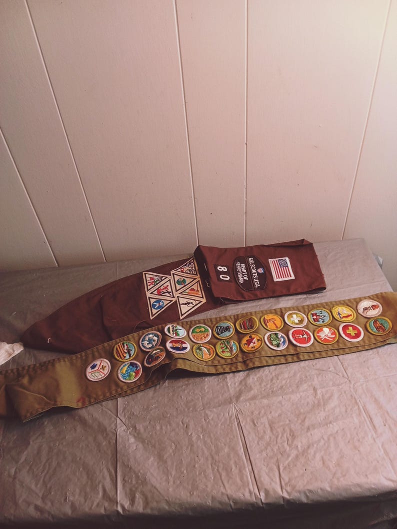 May include: A brown Boy Scouts sash with various merit badges and a brown neckerchief slide with merit badges. A brown bag with the text "SCOUTS USA" and an American flag is also present.