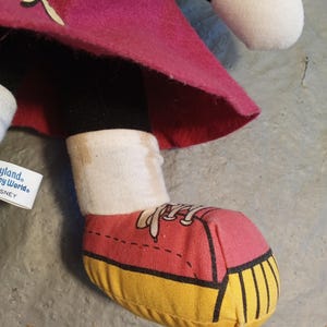 May include: A plush doll's foot wearing a red and yellow sneaker. The doll is wearing a pink skirt, black leggings, and a white sock. The doll's foot is on a gray surface. The doll is a Disney character.
