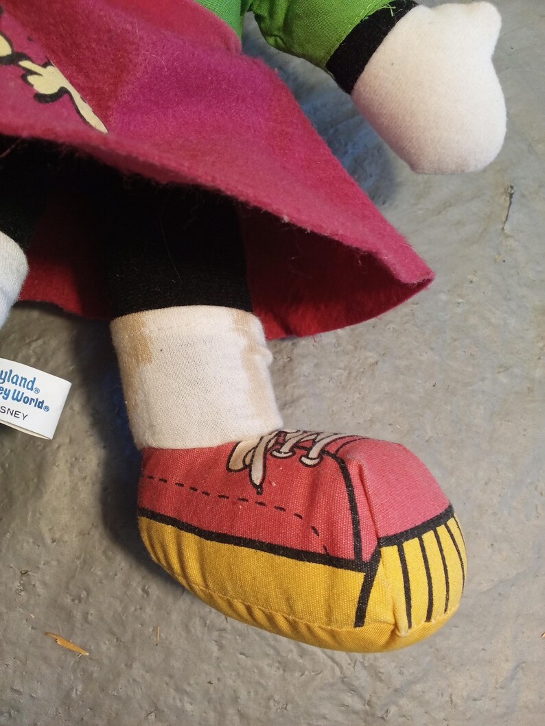 May include: A plush doll's foot wearing a red and yellow sneaker. The doll is wearing a pink skirt, black leggings, and a white sock. The doll's foot is on a gray surface. The doll is a Disney character.