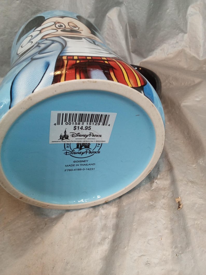 Pu&ograve; includere: Una tazza in ceramica Disney Parks con Topolino in un completo blu e rosso. La tazza ha un manico nero e una base azzurra. Il fondo della tazza presenta un logo e un testo Disney Parks.