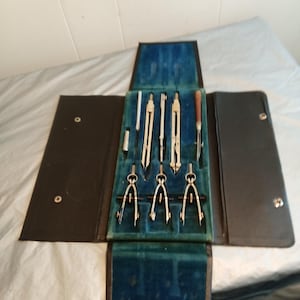 May include: A vintage drafting set in a black leather case. The set includes several compasses, dividers, and other drawing tools, arranged on a blue velvet-lined interior. The case is open, displaying the tools.