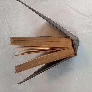 May include: An antique book with a worn, gray cover, partially open to reveal aged, yellowed pages. The spine is visible, and the pages are slightly fanned out, suggesting it has been well-read. The book's edges show signs of age.