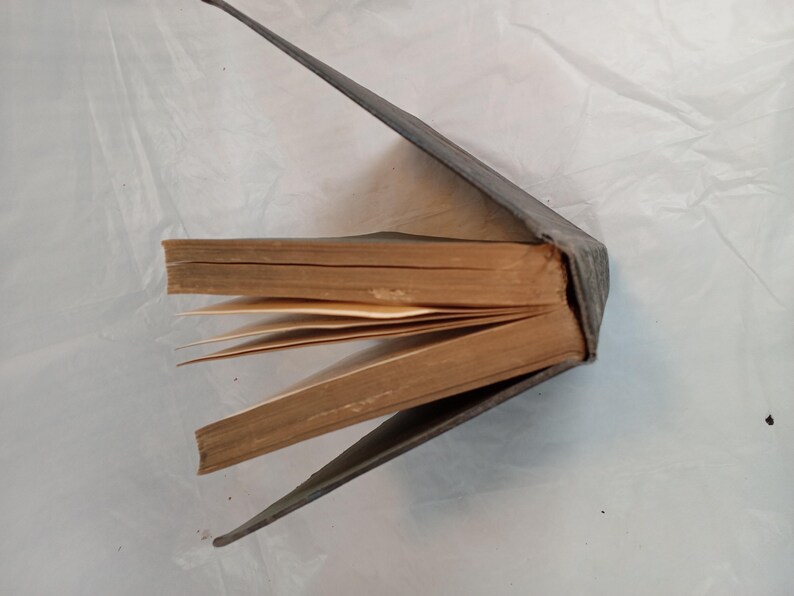 May include: An antique book with a worn, gray cover, partially open to reveal aged, yellowed pages. The spine is visible, and the pages are slightly fanned out, suggesting it has been well-read. The book's edges show signs of age.