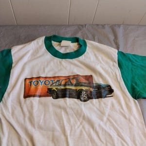 May include: White t-shirt with green sleeves and a vintage Toyota car graphic. The graphic features a black car with a yellow stripe and the word "TOYOTA" in a yellow box.