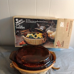 Puede incluir: Un plato de vidrio ámbar vintage con tapa, sentado dentro de una cesta de mimbre tejida. La caja dice "BRAVO Bakeware in a basket WFC-007G".