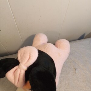 May include: A plush toy shaped like a cartoon character, featuring black ears, a pink bow, and a pink body. The toy has white accents on the face and feet. The toy is lying on a light-colored surface.