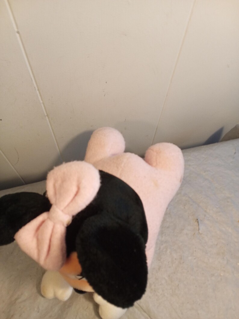 May include: A plush toy shaped like a cartoon character, featuring black ears, a pink bow, and a pink body. The toy has white accents on the face and feet. The toy is lying on a light-colored surface.