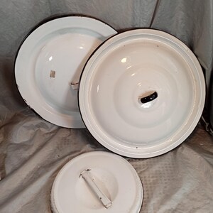 May include: Three white enamel pot lids with black trim. The lids are round and have a handle or knob on top. The lids are in various sizes and are arranged on a wrinkled, light gray surface. The lids appear to be vintage or antique.