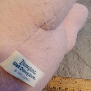 May include: A light pink plush toy with a tag that reads "Disneyland, Walt Disney World, &copy; 1984 DISNEY." A wooden ruler with inch markings is partially visible. The toy's texture appears soft and fuzzy.