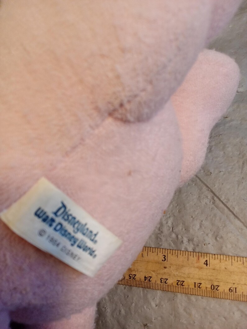 May include: A light pink plush toy with a tag that reads "Disneyland, Walt Disney World, &copy; 1984 DISNEY." A wooden ruler with inch markings is partially visible. The toy's texture appears soft and fuzzy.
