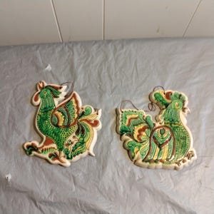 May include: Two ceramic rooster ornaments with green, yellow and brown painted details. The roosters are facing opposite directions and have a hole at the top for hanging.
