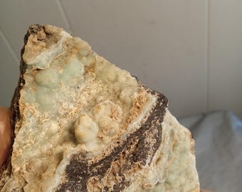 May include: A mineral specimen with a dark brown band and a pale green, bubbly surface. The rock has a beige, textured area and a jagged edge, showcasing natural geological formations.