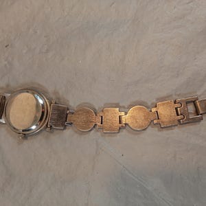May include: A silver-toned wristwatch featuring a round case and a link bracelet composed of alternating circular and rectangular metal segments. The watch has a plain, solid back casing and a small winding crown on the side of the watch head.