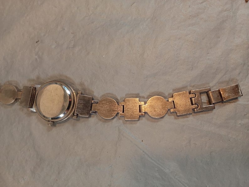 May include: A silver-toned wristwatch featuring a round case and a link bracelet composed of alternating circular and rectangular metal segments. The watch has a plain, solid back casing and a small winding crown on the side of the watch head.