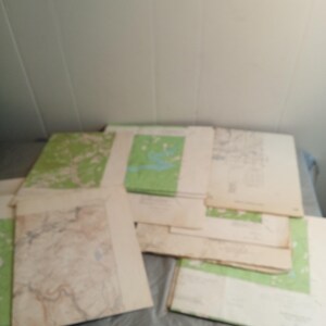 May include: A collection of vintage maps, featuring green and tan color schemes. The maps are stacked on top of each other, with some showing detailed geographical features. The paper has a slightly aged appearance.