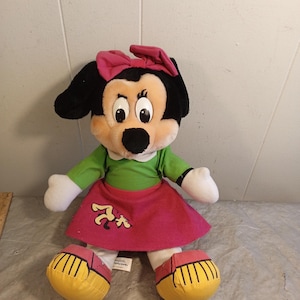May include: A plush Minnie Mouse doll with a black head, large eyes, and a pink bow. She wears a green shirt, a pink skirt with a cartoon graphic, and yellow shoes. The doll is a classic Disney character.