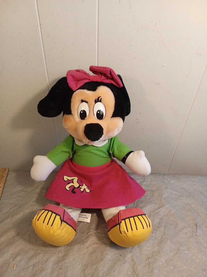 May include: A plush Minnie Mouse doll with a black head, large eyes, and a pink bow. She wears a green shirt, a pink skirt with a cartoon graphic, and yellow shoes. The doll is a classic Disney character.