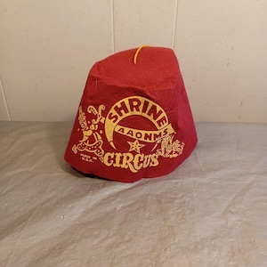 May include: A red felt fez hat with gold lettering and graphics. The hat features the words "Shrine Circus" in a circular design, with additional text and illustrations. A yellow string is attached at the top. Made in the USA.