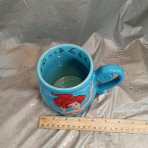 May include: A light blue ceramic mug featuring Ariel from The Little Mermaid. The mug has a curved handle and is decorated with images of Ariel and fish. The mug is approximately 4 inches tall.