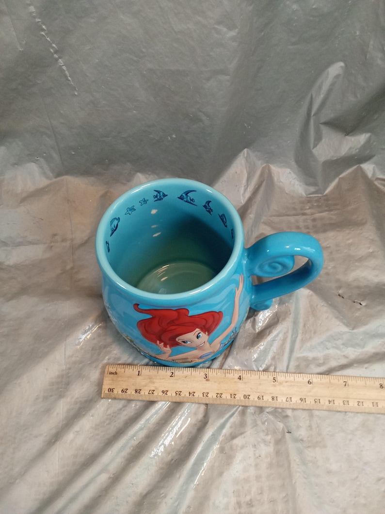 May include: A light blue ceramic mug featuring Ariel from The Little Mermaid. The mug has a curved handle and is decorated with images of Ariel and fish. The mug is approximately 4 inches tall.